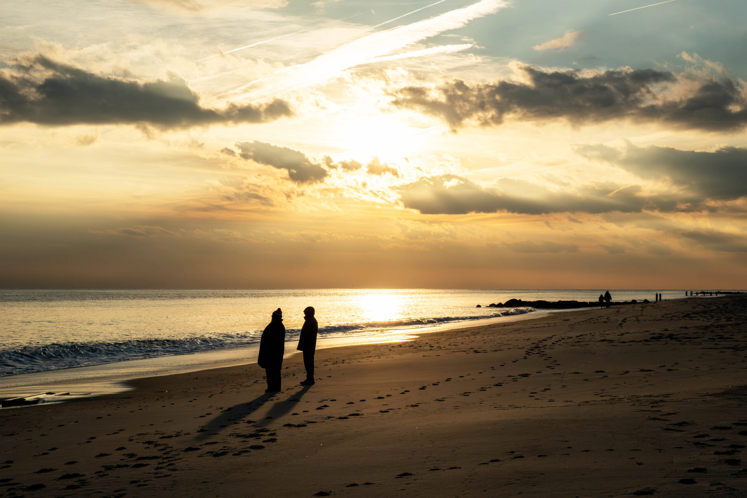 A wide view of the beach with the sun setting. Two people are standing on the beach silhouetted by the sun. There are some clouds in the sky making the sun hazy. The sky is orange, yellow, and blue. 