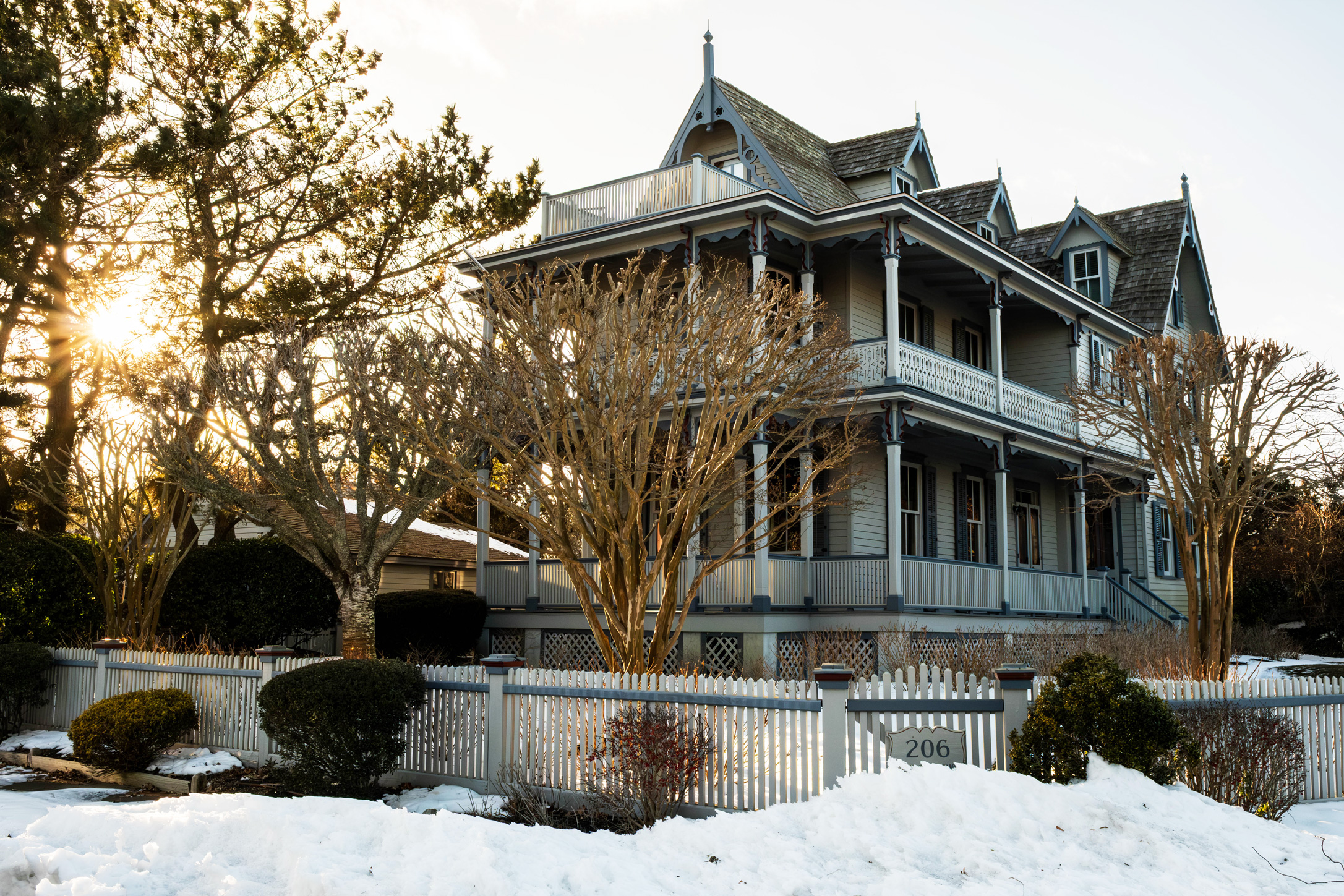 A wide view of a Victorian style home with snow on the ground and the sun shining behind trees.